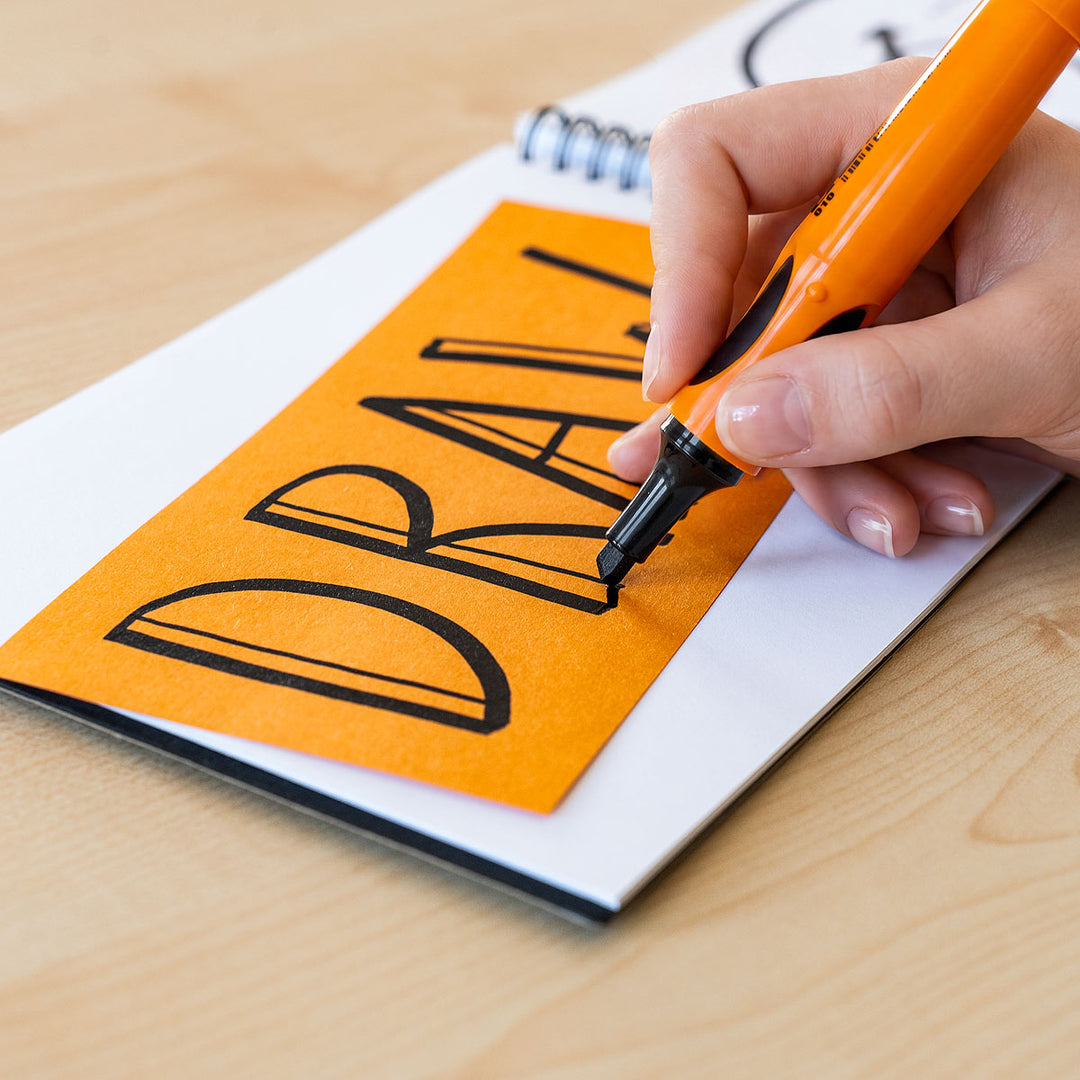 Hand holding an orange Neuland marker with black nib writing on a piece of paper with 'DRAW' text.