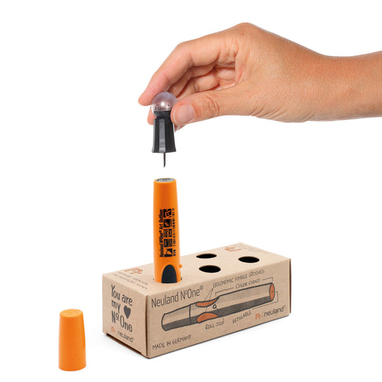 Hand holding an orange and black pen over a cardboard refill box with 'Neuland' branding.