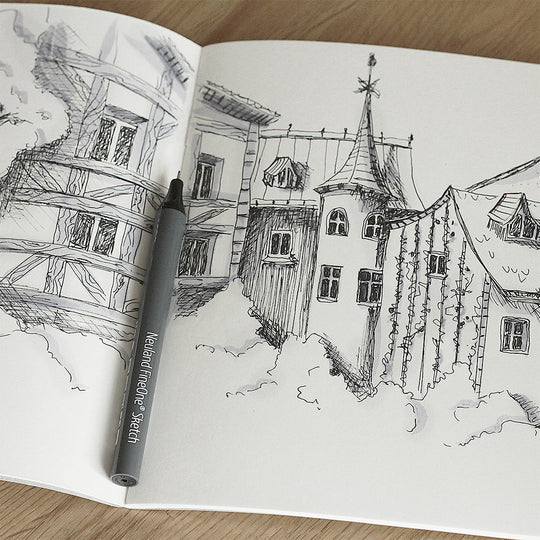 Open Neuland notebook with a black and white drawing of a village scene on a wooden surface using FineOne Sketch pens.