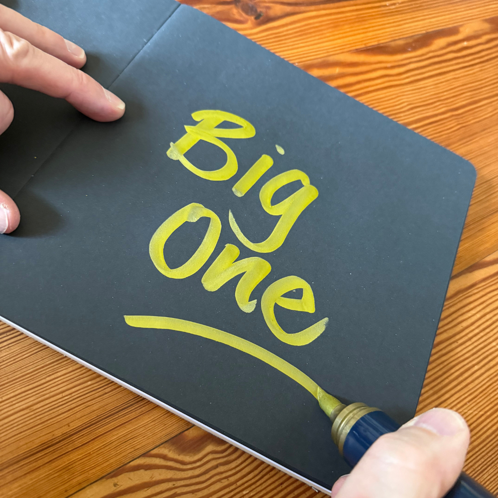 Handwritten 'Big One' on a black notebook with a Neuland gold metallic brush nib marker, on a wooden surface.