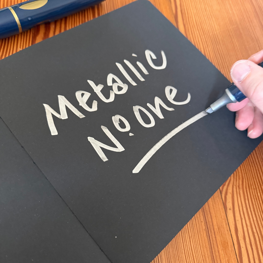 Handwriting on black paper with a Neuland pen labelled 'Metallic No. One' on a wooden surface.